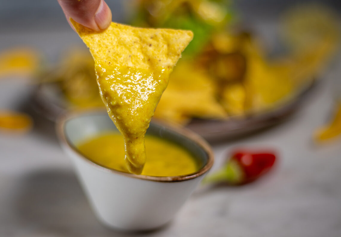 A Tortilla, being dipped into a vegan cheese dip. The cheese dip drops stringy from the tortilla.